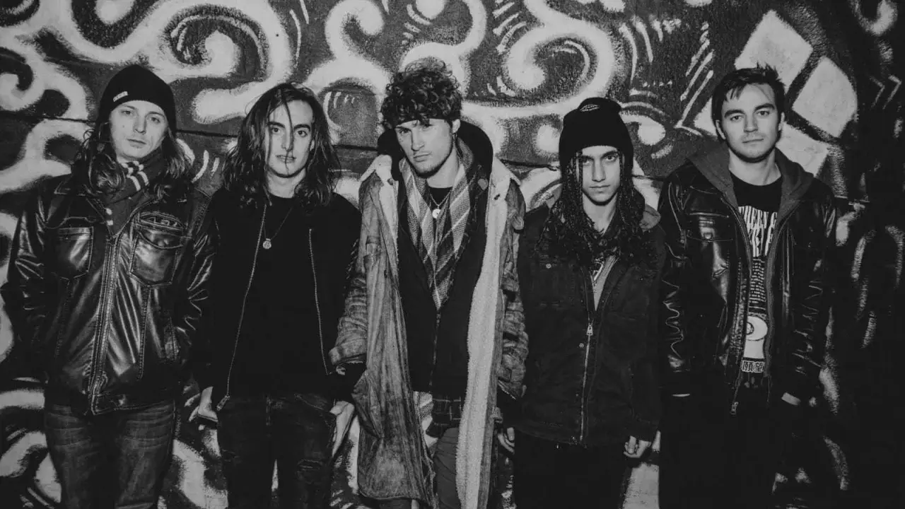 Fem unga män i rockkläder poserar framför en graffiti-vägg. De ser ut som ett band från 90 tals rock.