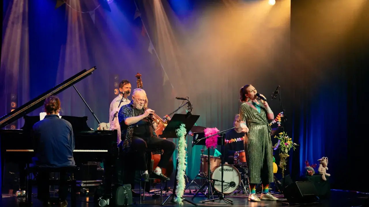 En jazzgrupp uppträder på scenen under Stockholm Jazz Festival. En sångerska i glittrig klänning sjunger, en man spelar trumpet, en annan står vid basen och en person sitter vid pianot.