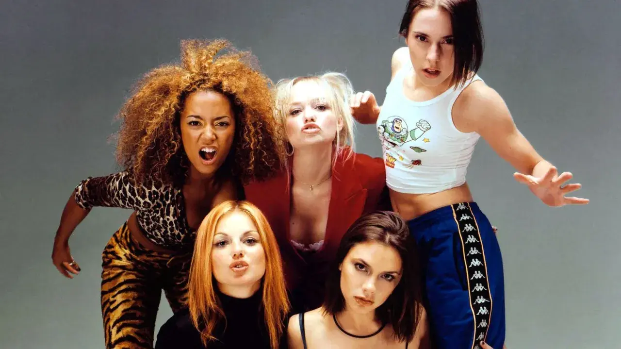 Medlemmar av Spice Girls poserar energiskt. Mel B i leopardmönster, Emma B i rött, Mel C i Buzz Lightyear-linne, Geri H med rött hår och Victoria B med mörkt hår.