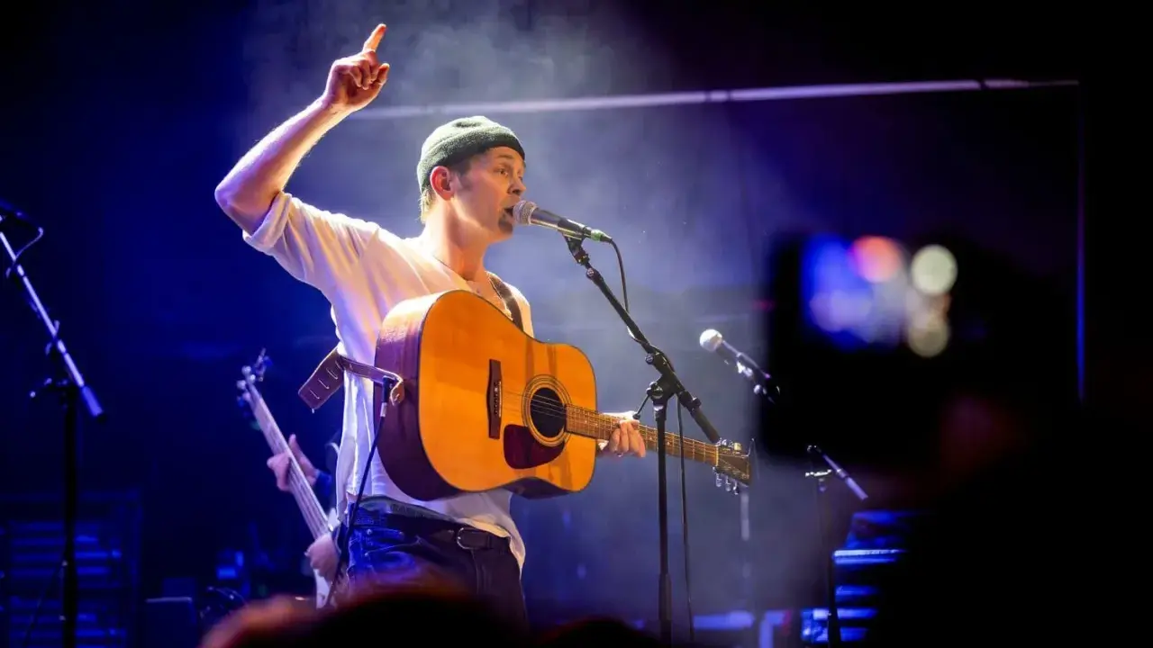 Joel Alme konsert. Sångaren med gitarr och mössa pekar uppåt under en blåbelyst spelning.