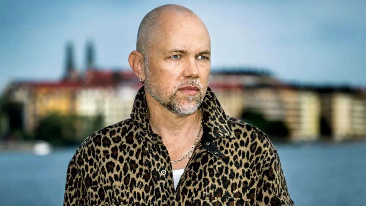 Peter LeMarc i leopardmönstrad jacka, redo för en konsert. Bakgrunden visar en stad vid vattnet.