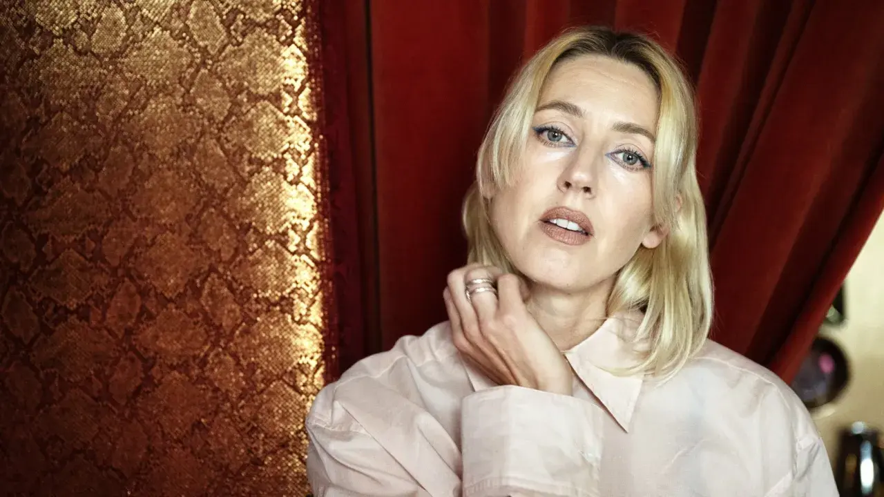 Veronica Maggio ung, med blont hår och blå eyeliner, poserar framför en gyllene ormskinnsmönstrad bakgrund och röda draperier.