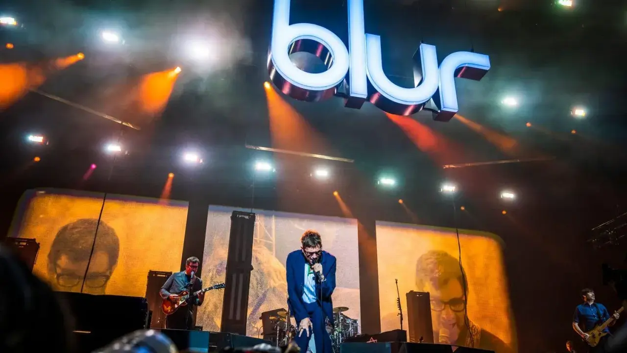 Medlemmar av Blur uppträder på scenen under en konsert, med den ikoniska logotypen lysande ovanför.