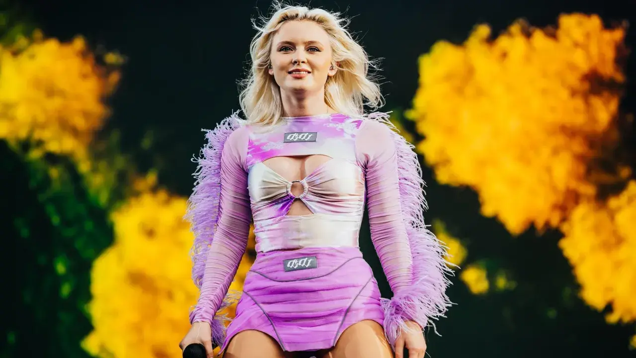 Zara Larsson konsert. Hon står på scenen i en lila och rosa outfit med fjädrar på ärmarna.