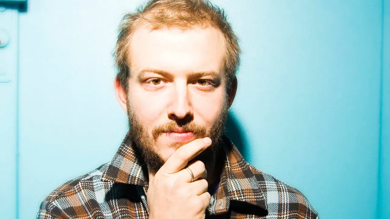 Bon Iver Sverige: en man med skägg och flanellskjorta funderar mot en turkos bakgrund.