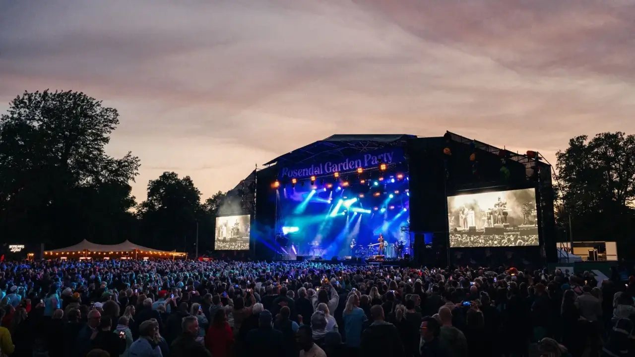 Stor publik vid Rosendal Garden Party. En scen med blå ljus och artister, omgiven av träd och en vacker kvällshimmel.