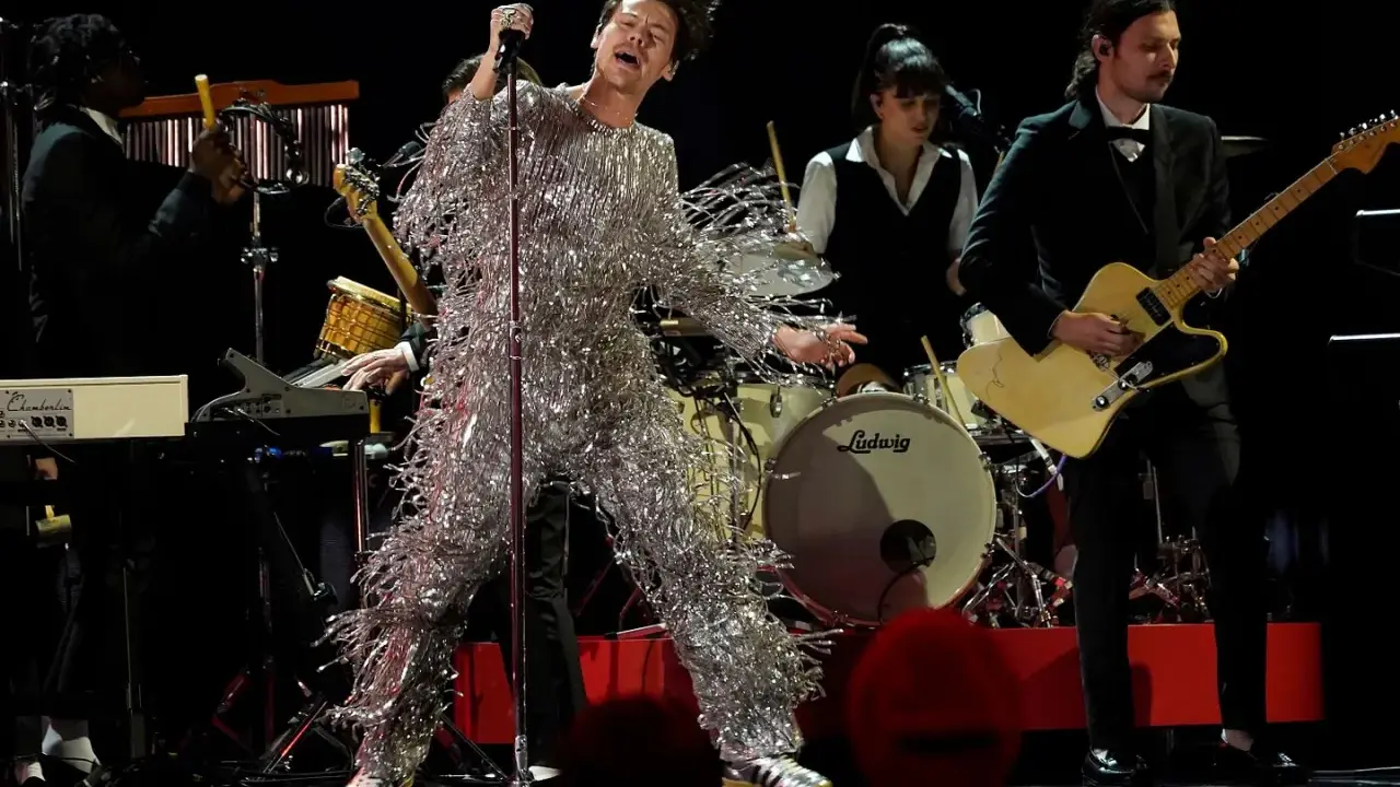 Harry Styles i glittrig outfit på en magisk harry styles konsert. Bandet spelar bakom honom, en gitarrist i kostym och en trummis.