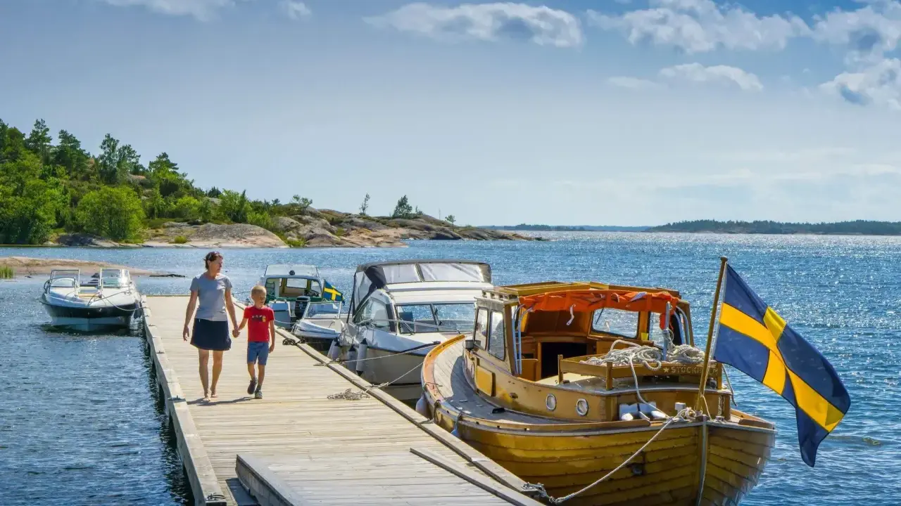 En kvinna och ett barn går på en brygga med båtar och en svensk flagga. En perfekt bild av en semester i Sverige.