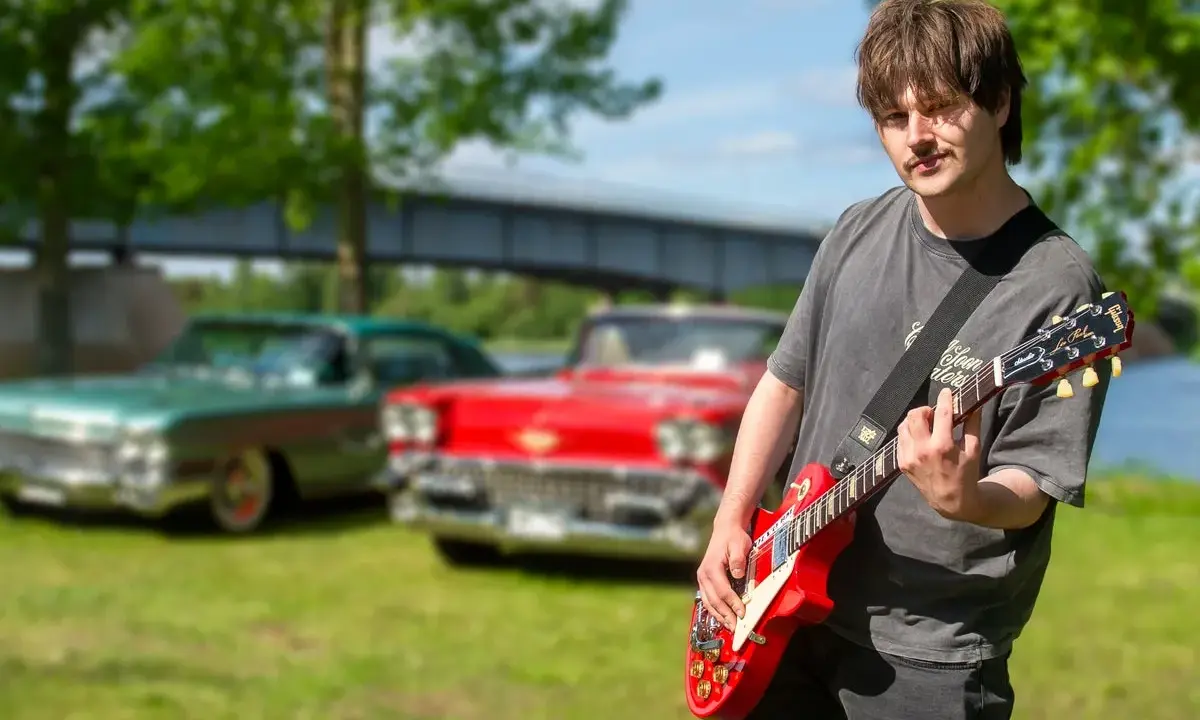 Erik Meduza spelar gitarr framför klassiska bilar vid en sjö.