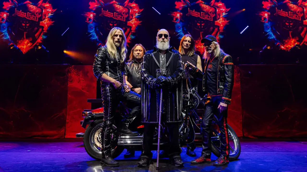 Medlemmar av Judas Priest poserar på scenen med en motorcykel. Bandets logotyp syns i bakgrunden.