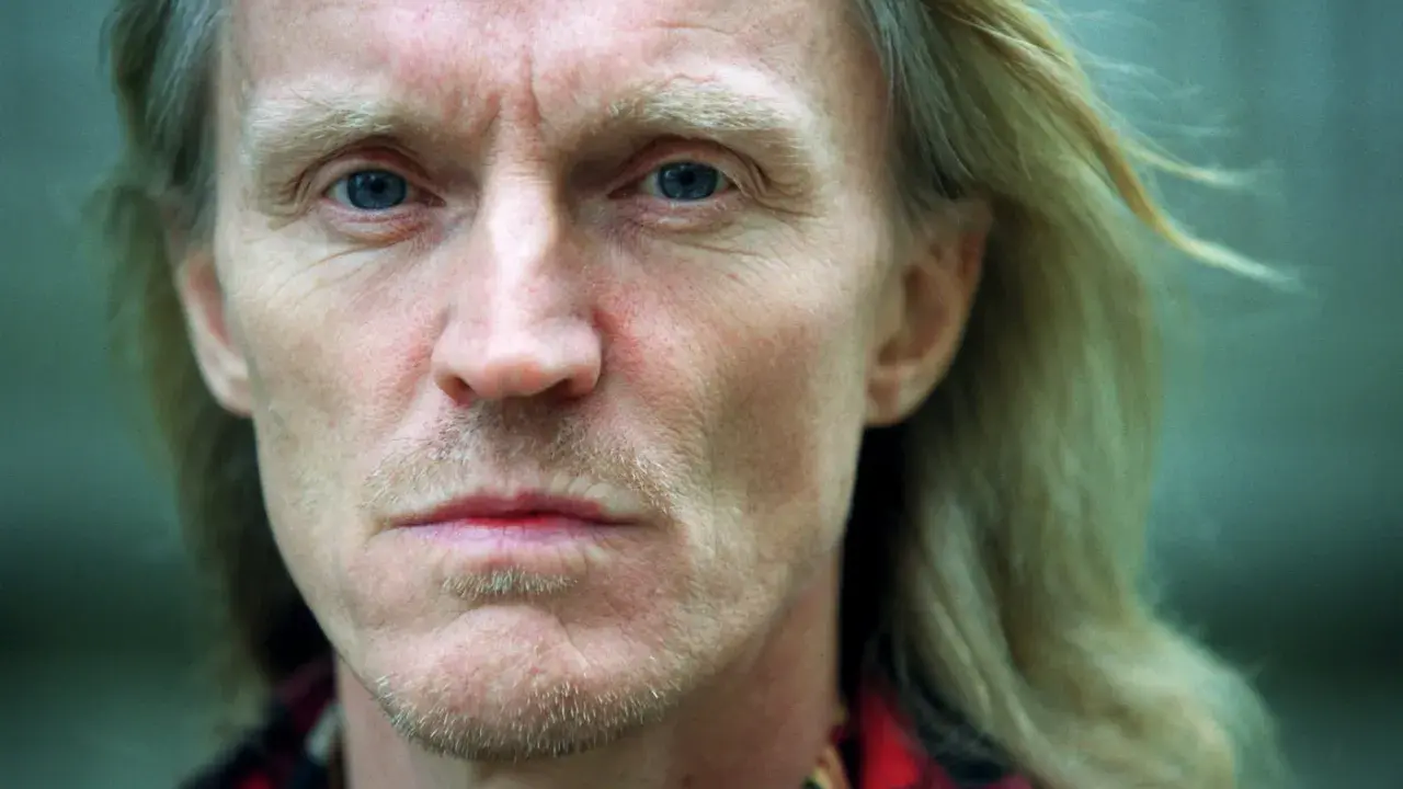 Närbild på John Holm med blå ögon och blont hår. Hans ansikte bär spår av livserfarenhet, en sordinerad blick.