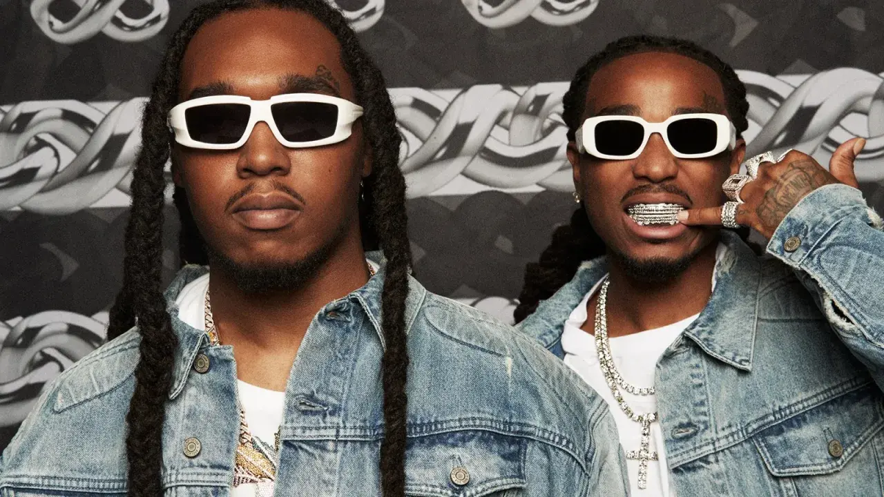 Två medlemmar av Migos, klädda i denimjackor och vita solglasögon, poserar med bling och grillz.