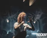 Lorna Shore Sverige: Sångaren med flammigt hår och tatueringar greppar mikrofonen på scenen, omgiven av rök och dramatiskt ljus.