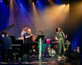 En jazzgrupp uppträder på scenen under Stockholm Jazz Festival. En sångerska i glittrig klänning sjunger, en man spelar trumpet, en annan står vid basen och en person sitter vid pianot.