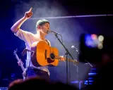 Joel Alme konsert. Sångaren med gitarr och mössa pekar uppåt under en blåbelyst spelning.