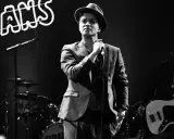 Bruno Mars i Sverige, med hatt och kavaj, sjunger i mikrofonen. Bakom honom syns ett band och neonljus.