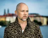 Peter LeMarc i leopardmönstrad jacka, redo för en konsert. Bakgrunden visar en stad vid vattnet.