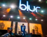 Medlemmar av Blur uppträder på scenen under en konsert, med den ikoniska logotypen lysande ovanför.