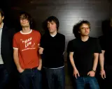 Medlemmar av Radiohead står uppradade framför en mörk trävägg. Thom Yorke bär solglasögon.