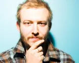 Bon Iver Sverige: en man med skägg och flanellskjorta funderar mot en turkos bakgrund.