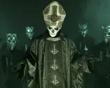 Papa Emeritus IV, klädd i en detaljerad mörkgrön prästrock med korsmotiv, står framför en grupp maskerade följeslagare med horn.