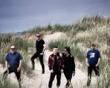 Fem män poserar i sanddyner med högt gräs. De ser ut att njuta av gyllene tider unga, med en känsla av tidlöshet.