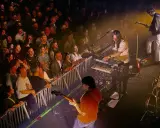 Publik njuter av Altin Gün i Stockholm. En gitarrist spelar, en keyboardist bakom Roland-syntar, och en entusiastisk publik bakom stängsel.