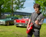 Erik Meduza spelar gitarr framför klassiska bilar vid en sjö.