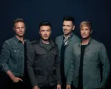 Medlemmar av Westlife poserar för en studiofotografering. De bär moderna jackor och ser direkt in i kameran.