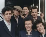 Medlemmar av the Pogues poserar glatt utomhus, några med cigaretter.