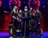 Medlemmar av Judas Priest poserar på scenen med en motorcykel. Bandets logotyp syns i bakgrunden.