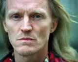 Närbild på John Holm med blå ögon och blont hår. Hans ansikte bär spår av livserfarenhet, en sordinerad blick.