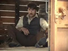 Brandon Sklenar sitter i ett stall, klädd i väst och skjorta, med en mustasch och ett sår i ansiktet.
