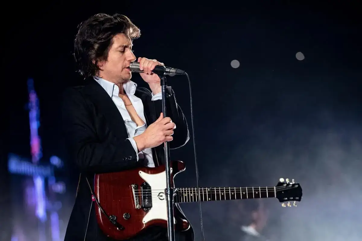 Alex Turner sjunger i mikrofonen med sin gitarr i handen, iklädd en mörk kavaj och ljus skjorta.