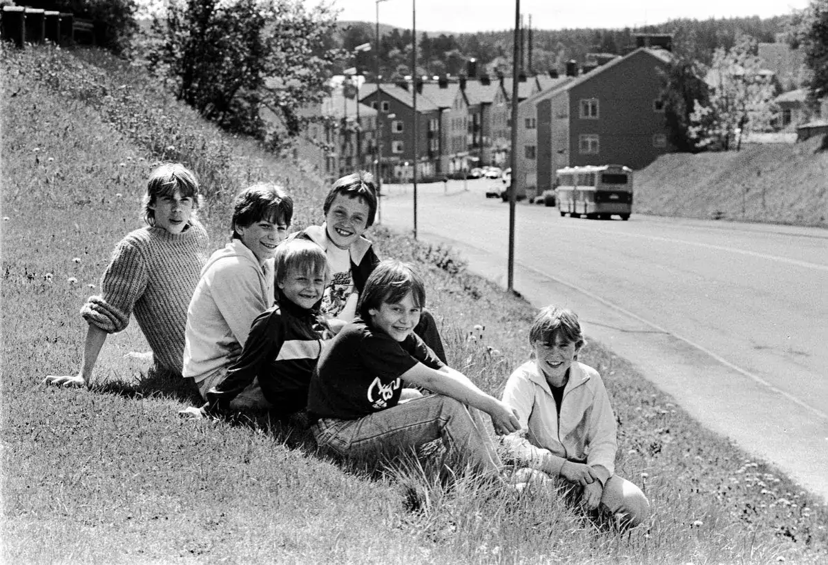 Sju glada barn, the pinks medlemmar, sitter i gräset en solig dag. I bakgrunden syns hus och en buss.