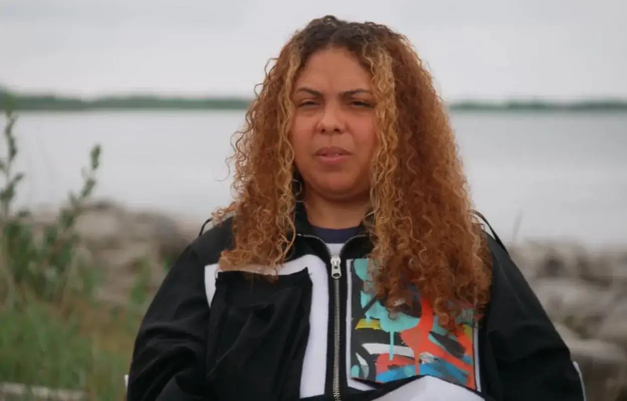 En kvinna med lockigt, rödbrunt hår och en jacka med konstnärligt tryck, en sann mapei artist, sitter utomhus vid vattnet.