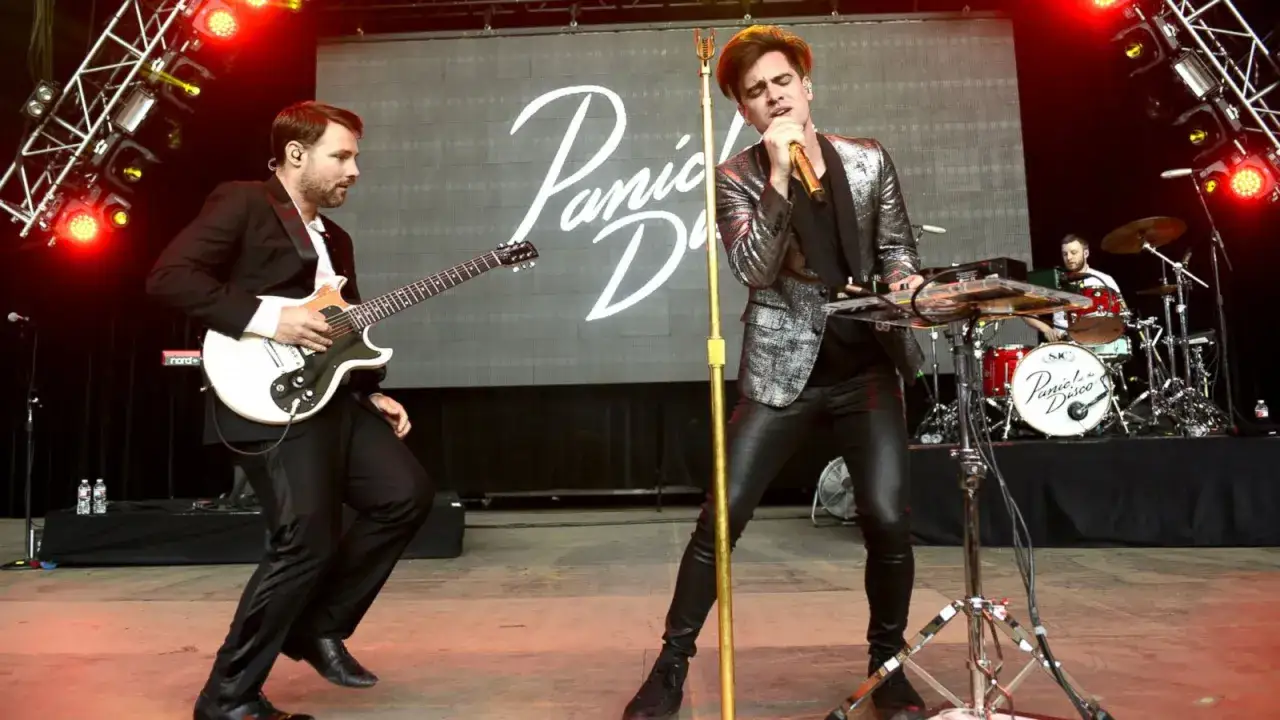 Medlemmar av Panic! at the Disco uppträder live. Sångaren i glittrig kavaj och läderbyxor sjunger i en gul mikrofon. Gitarristen i kostym spelar gitarr.