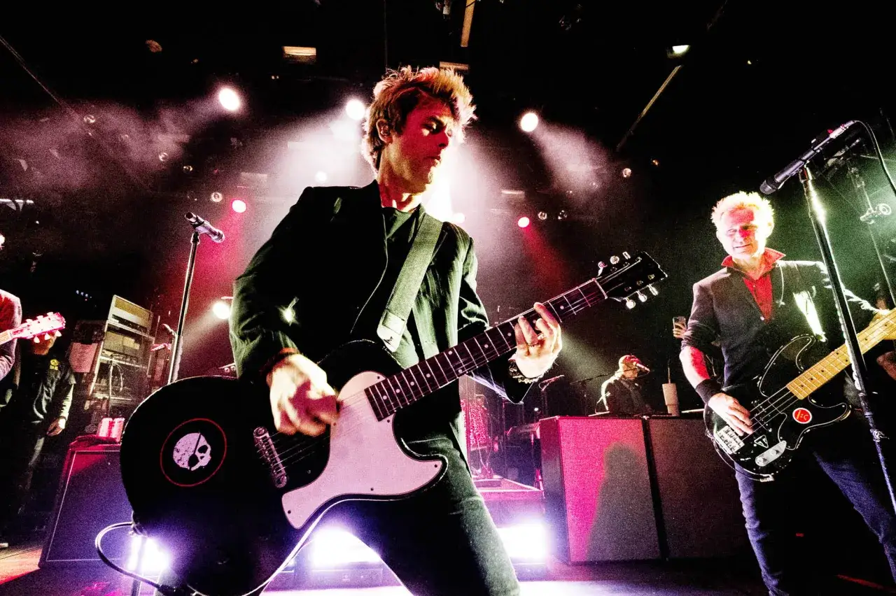 Medlemmar av Green Day spelar gitarr och bas på scenen, upplysta av starka strålkastare och rök.