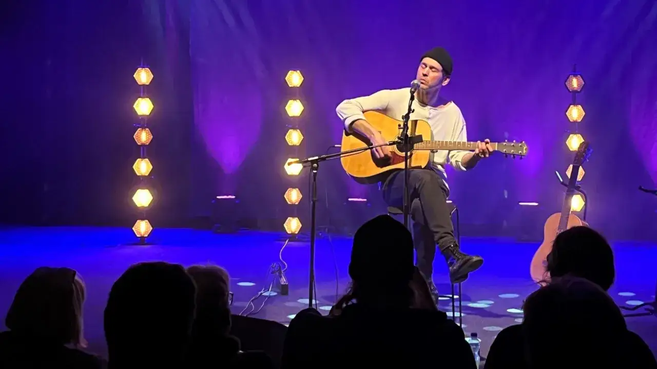 Joel Alme uppträder i Stockholm, sittande med gitarr på en scen med lila ljus och dekorativa lampor.