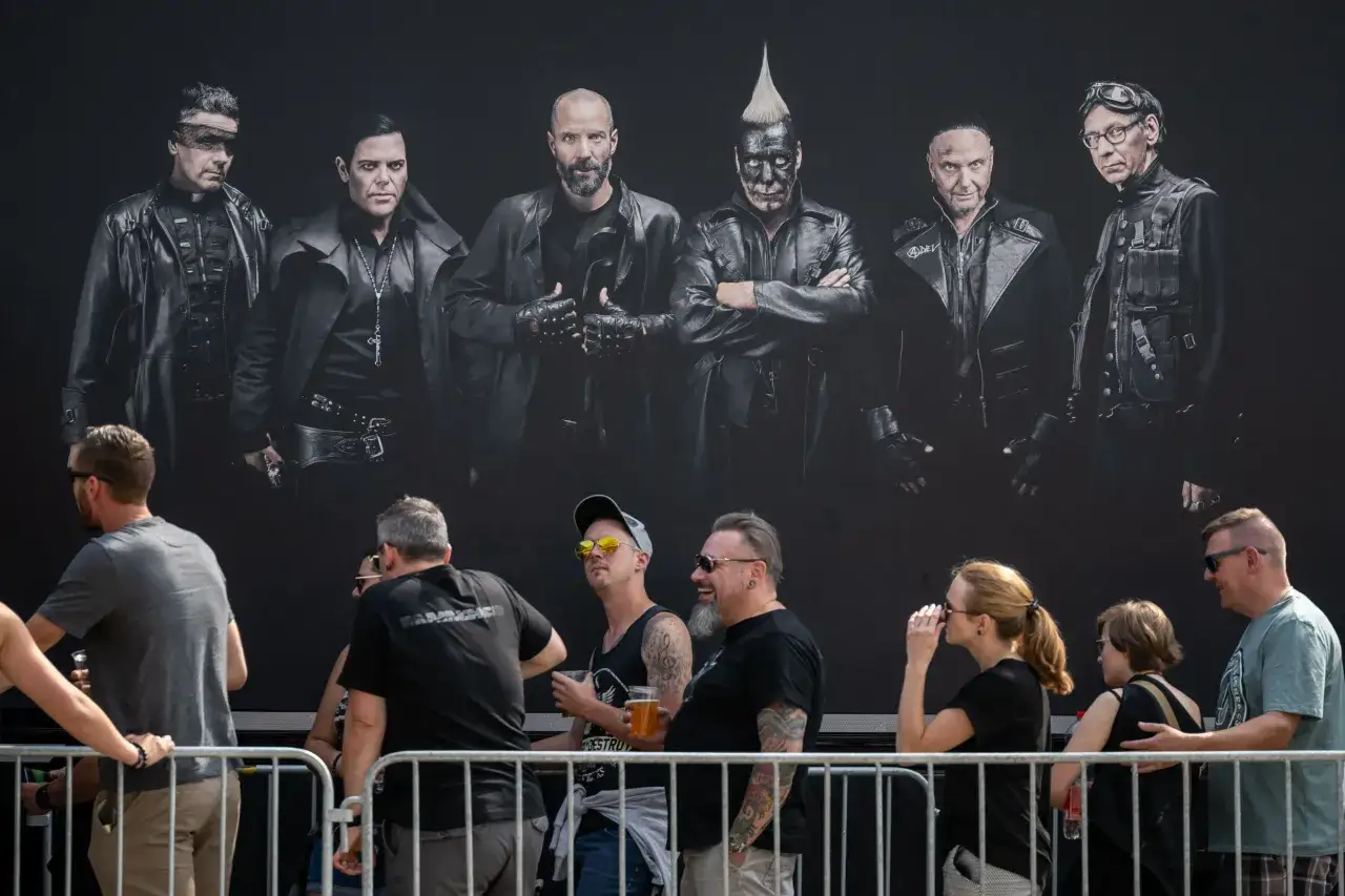 Medlemmar av Rammstein poserar i sina karaktäristiska scenkläder framför en mörk bakgrund. Nedanför syns en publik som väntar.