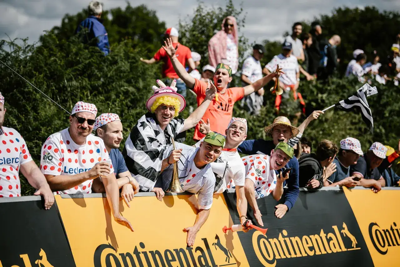 Glada åskådare med prickiga tröjor och hattar hejar på Tour de France. En man spelar trumpet.