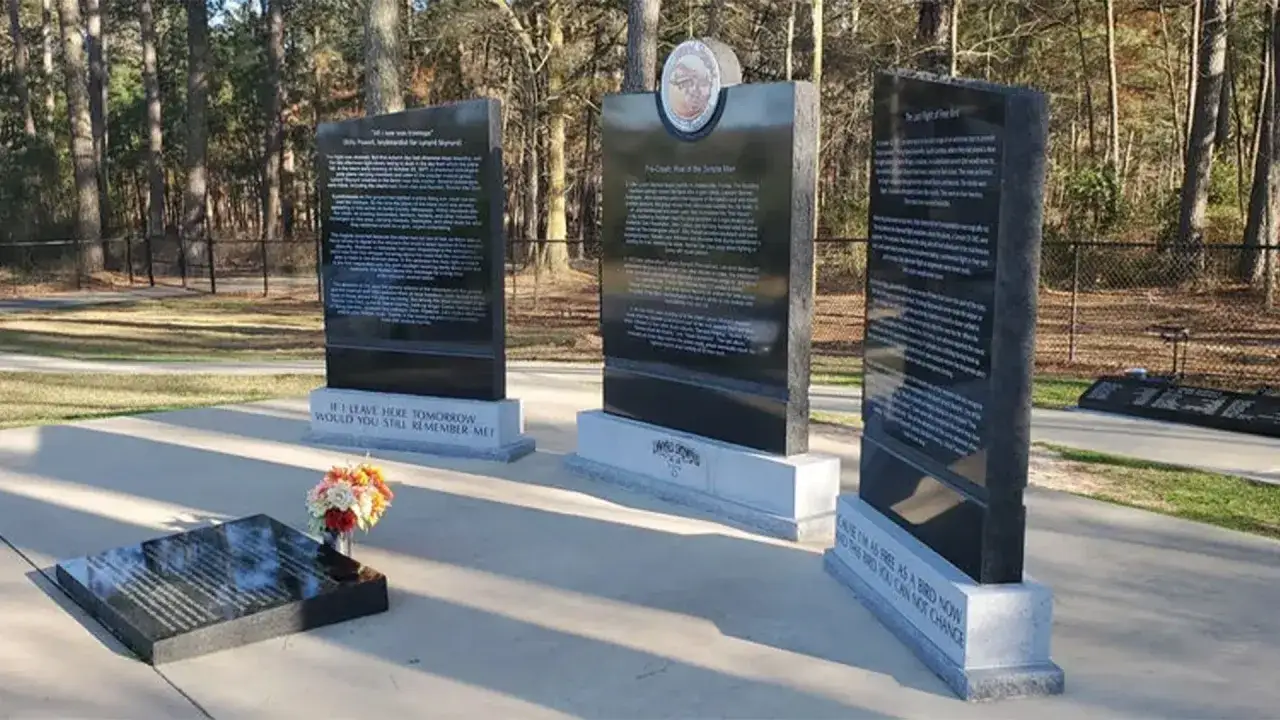 Minnesmärke över lynyrd skynyrd flygolycka. Tre stora svarta stenar med text och en mindre gravsten med blommor.