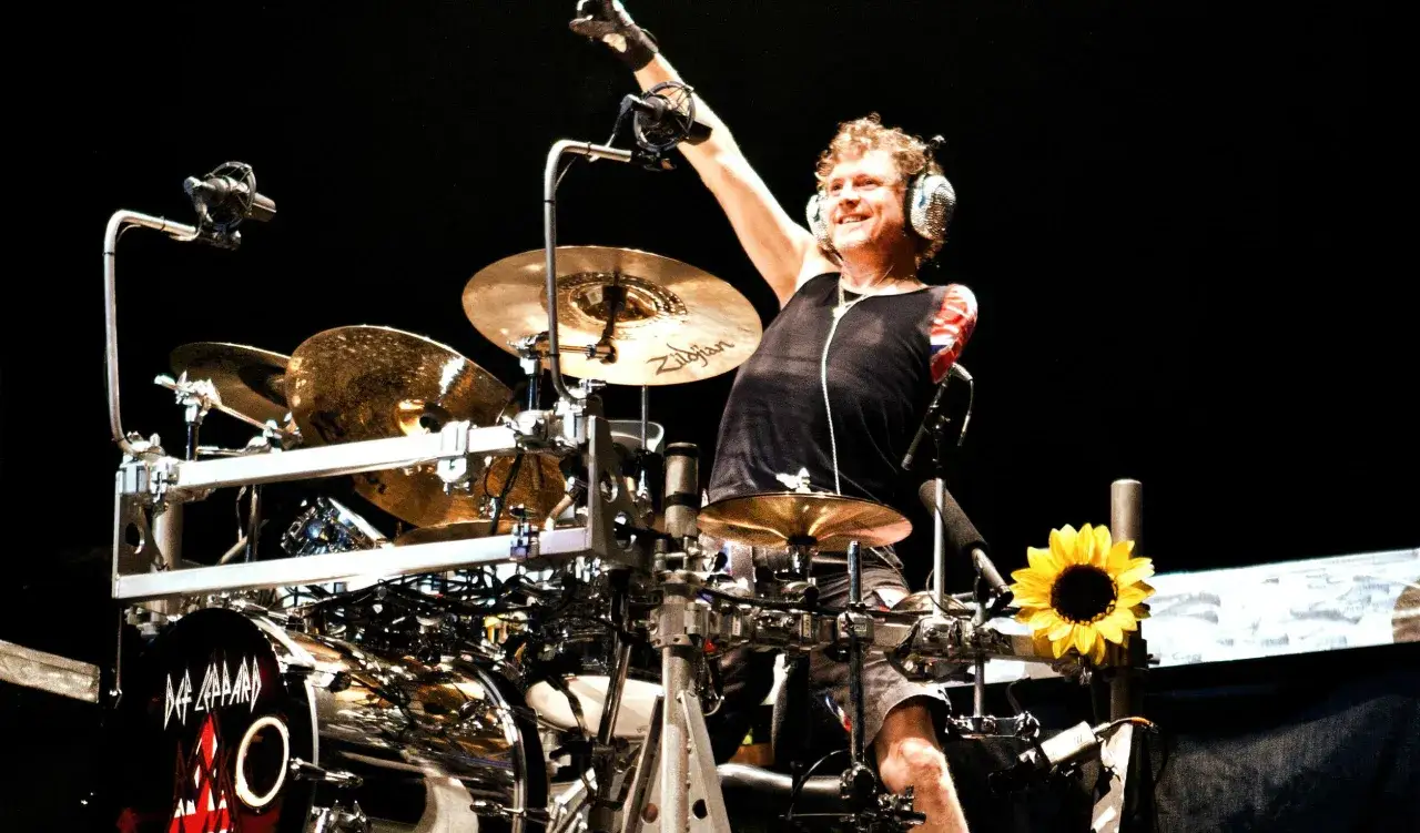 Rick Allen, Def Leppard trummis, höjer handen i luften vid sitt trumset. En solros står bredvid.