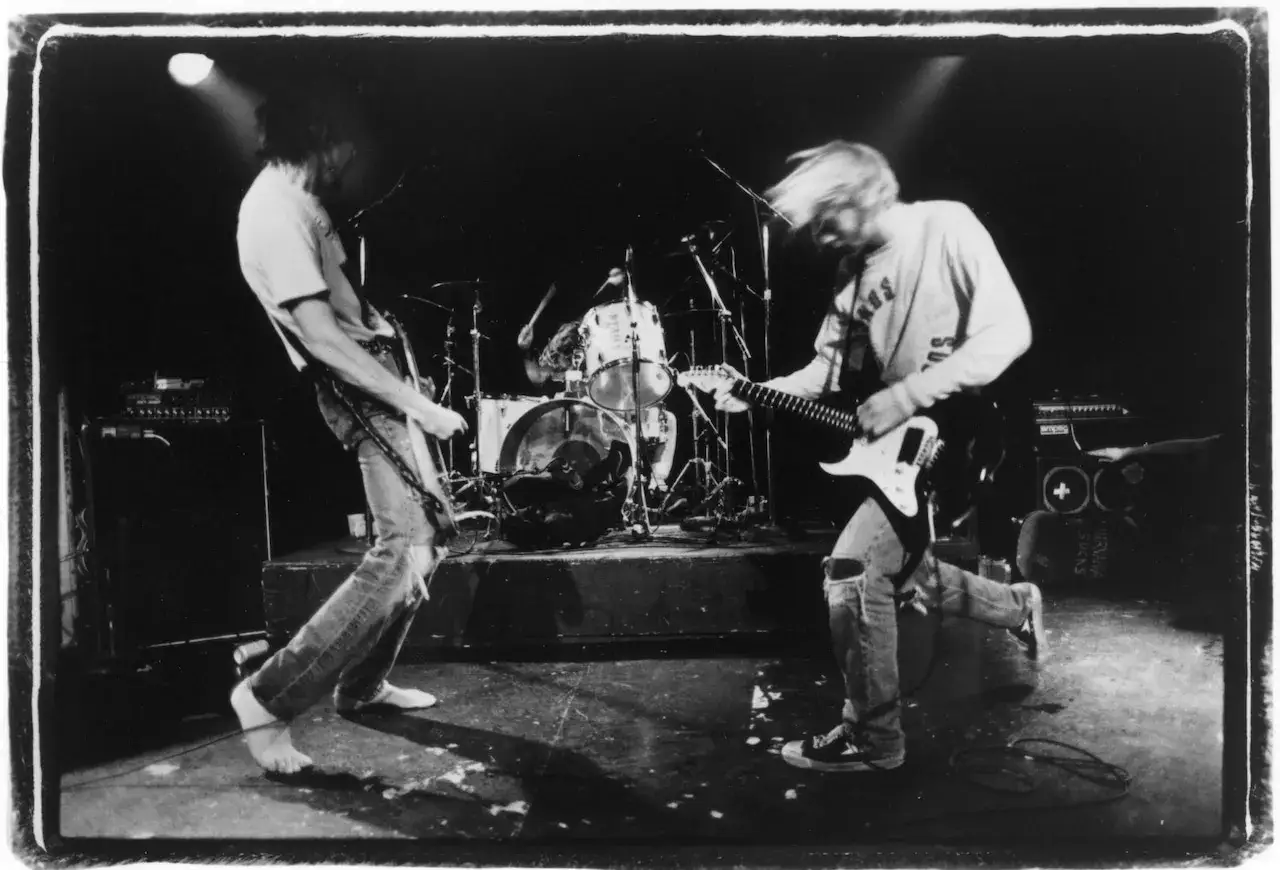 Scen från en 90 tals rockkonsert. En gitarrist med blont hår spelar intensivt, medan en annan gitarrist med mörkt hår står till vänster. Trummisen syns i bakgrunden.