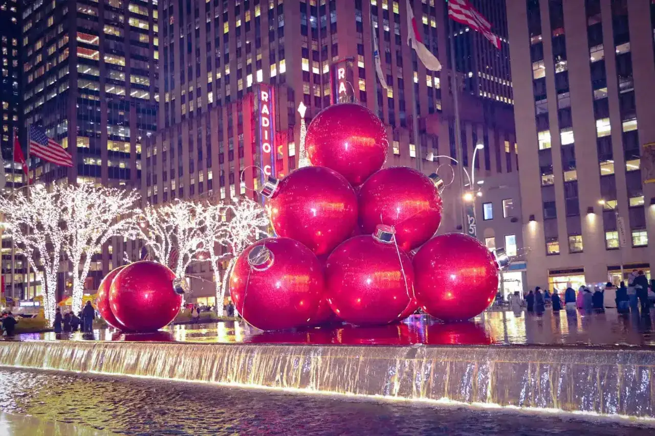 Stora röda julgranskulor glittrar vid en fontän i New York. Julbelysning lyser upp träden och skyskraporna i bakgrunden.