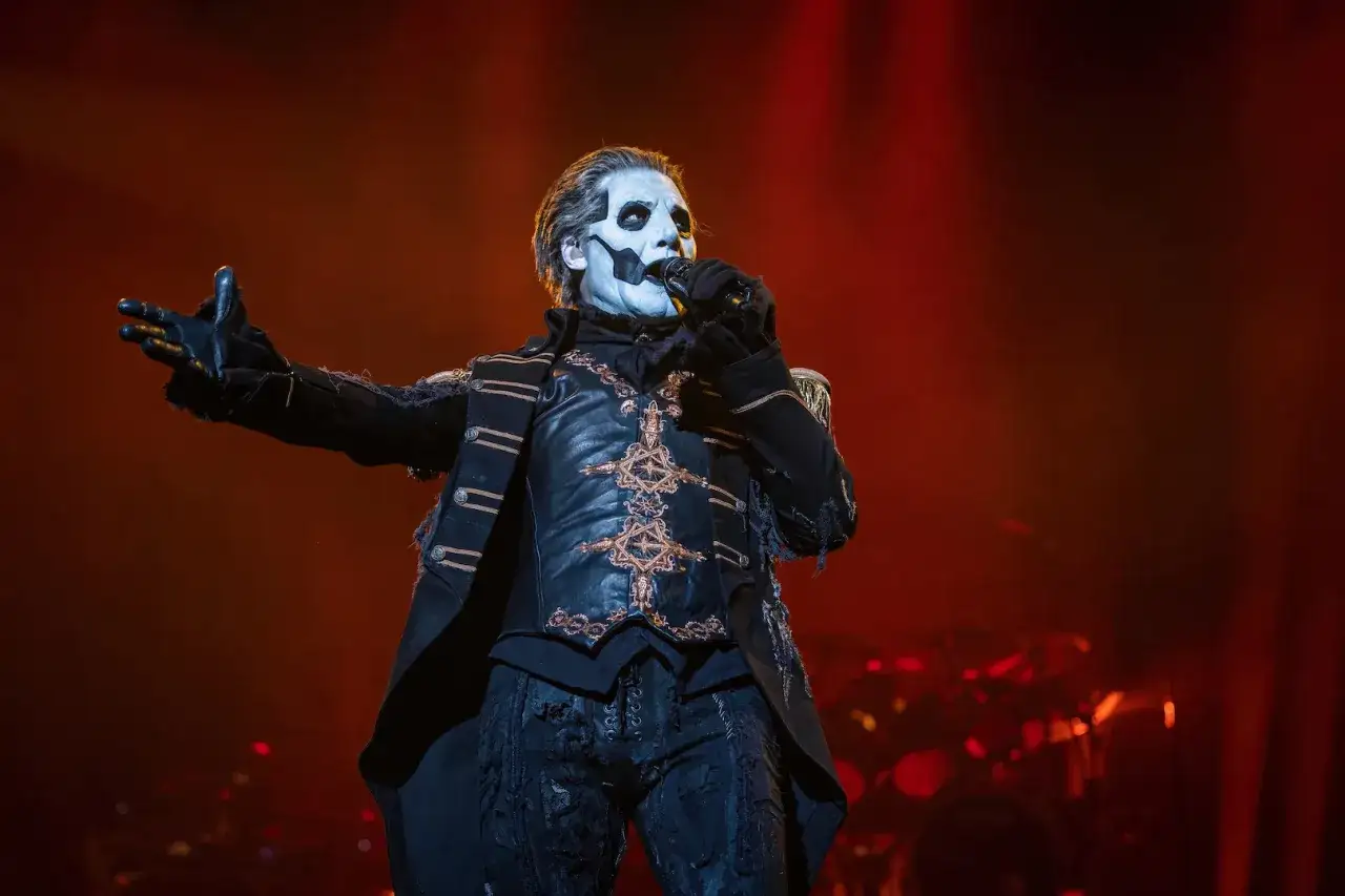 Papa Emeritus IV, med vit ansiktsmålning och i en detaljerad, mörk scenkostym, sjunger med mikrofonen i handen.