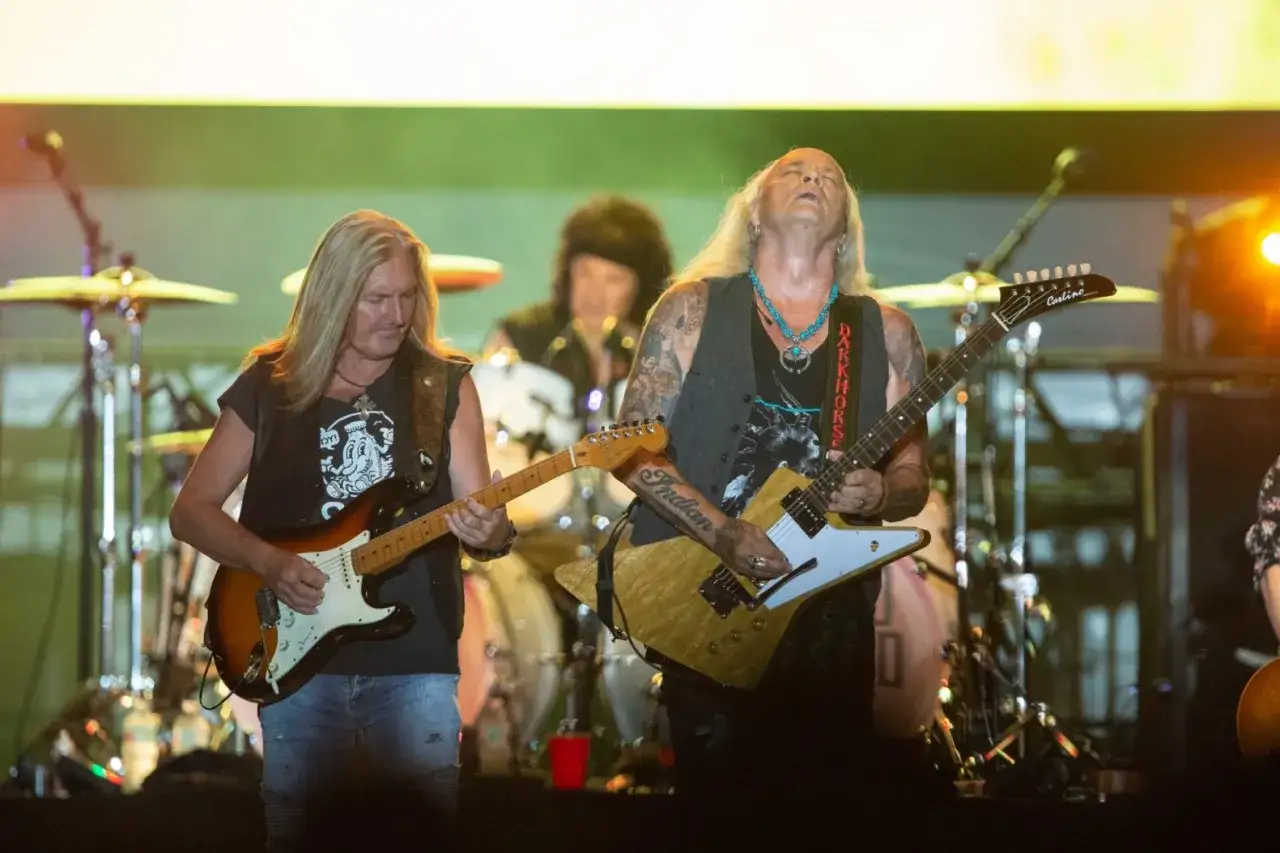Medlemmar av Lynyrd Skynyrd spelar gitarr på scenen. En trummis syns i bakgrunden.