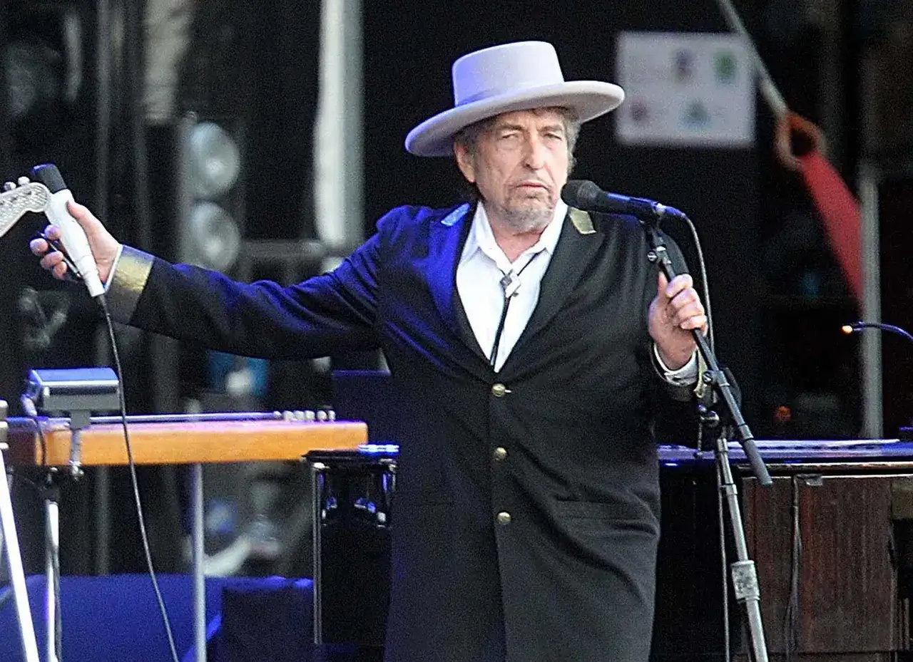 Bob Dylan med hatt och kavaj på scen i Stockholm, håller mikrofon och gitarr.