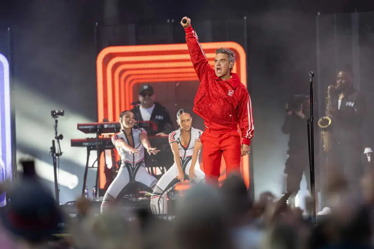 Robbie Williams i Sverige, glittrig röd jacka, mikrofon i luften, dansare och musiker på scenen.