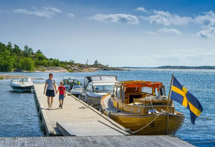 En kvinna och ett barn går på en brygga med båtar och en svensk flagga. En perfekt bild av en semester i Sverige.